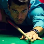 A young Caucasian man with short dark hair wearing a teal shirt, leaning over a pool table and lining up a shot